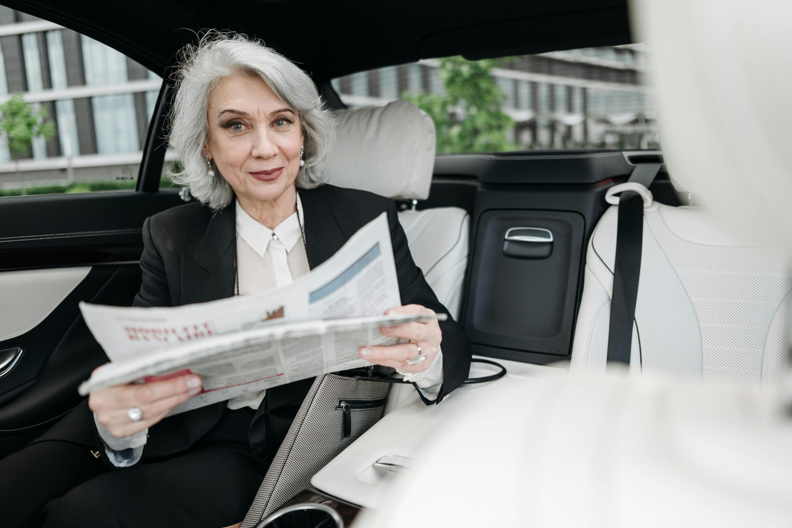 Senior businesswoman reading a newspaper while seated in a car, smiling confidently.